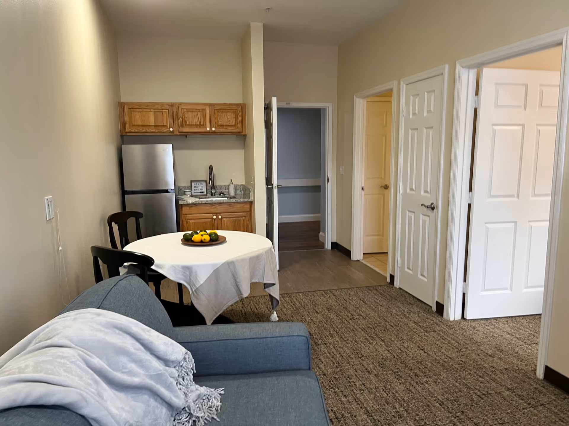 Interior view of a small assisted living apartment with a blue couch covered by a light gray blanket in the foreground. A round dining table with a white tablecloth and a bowl of fruit is next to two wooden chairs. In the background, there is a kitchenette with wooden cabinets, a stainless steel refrigerator, a sink, and a small framed sign. Three white doors lead to other rooms, and the floor is carpeted with a beige pattern.