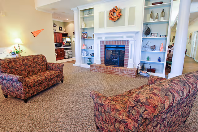 A cozy living room area with two floral patterned sofas facing a white fireplace with a brick hearth. The fireplace is flanked by built-in shelves decorated with various vases, framed pictures, and decorative items. A fall-themed wreath hangs above the fireplace. The room has beige walls and carpeted flooring, with a glimpse of a kitchen area in the background.