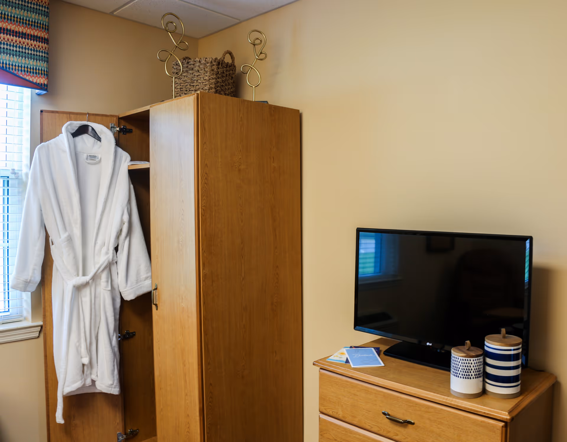 A bedroom corner with an open wooden wardrobe holding a white bathrobe, a dresser with a flat-screen TV and decorative jars, and a woven basket on top of the wardrobe.