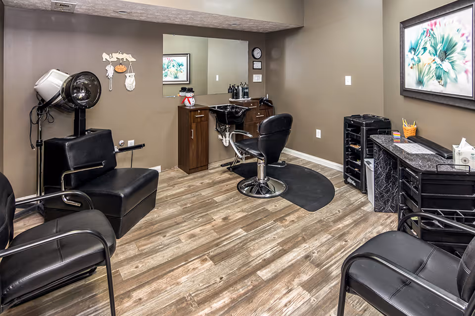 Interior of a hair salon area in an assisted living facility featuring black salon chairs, a hair dryer, a wash basin with a chair, a large mirror, wooden cabinets, and a framed floral painting on the wall. The floor is wood-patterned and the walls are painted beige.
