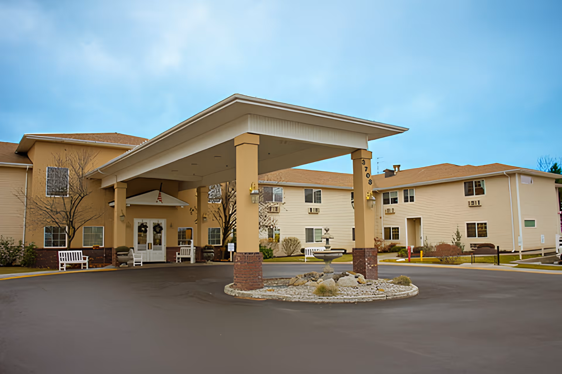 Exterior view of Avamere at South Hill senior living facility showing a covered entrance with pillars, a circular driveway, benches, and a small fountain in the center of the driveway. The building is two stories with beige walls and a brown roof under a blue sky.