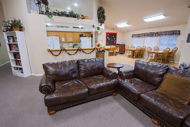 Interior view of a senior living facility common area featuring two brown leather sofas arranged in an L-shape on a carpeted floor. Behind the sofas is a kitchen area with wooden cabinets, white appliances, and a pass-through window decorated with garland. To the right, there is a dining area with wooden tables and chairs, a bulletin board on the wall, and windows with valance curtains allowing natural light.