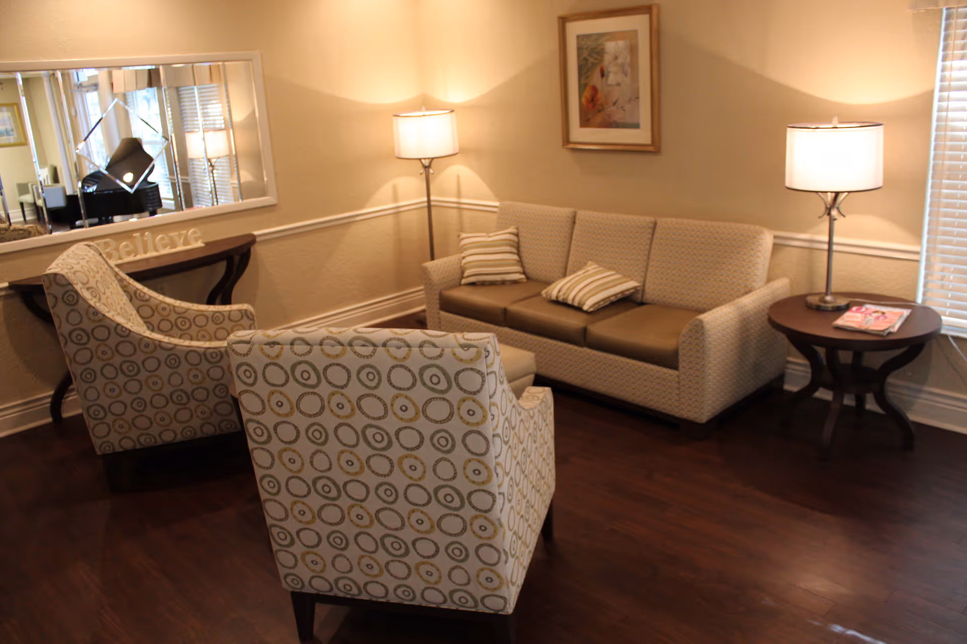Warmly lit lounge with a sofa, two patterned armchairs, side tables, lamps, and a wall mirror.