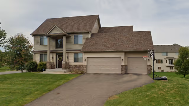 A two-story suburban house with a brown roof and beige siding, featuring a three-car garage and a basketball hoop on the driveway. The house is surrounded by a well-maintained lawn and some trees.