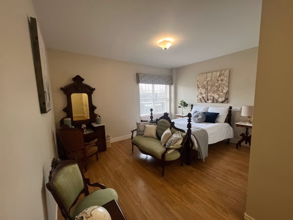 A cozy bedroom with wooden flooring featuring a bed with white linens and decorative pillows, a vintage-style green loveseat with multiple cushions, a wooden vanity with a mirror and chair, a green upholstered chair, a window with blinds and a floral valance, and a floral painting on the wall above the bed.