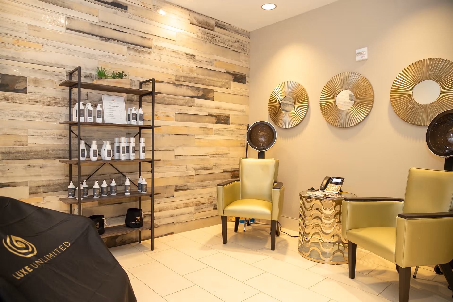 Interior of a salon area with two yellow salon chairs under hair dryers, a small metallic side table with a telephone, a wooden wall with a black metal shelf holding various hair care products, and three decorative round mirrors on the wall.