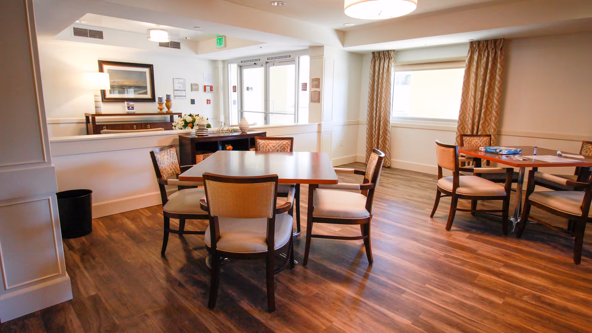 A well-lit common area with wooden flooring featuring two square tables surrounded by cushioned chairs. The room has large windows with patterned curtains, a sideboard with decorative items and a framed picture on the wall, and an exit door with glass panels in the background.