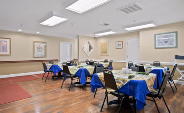 A dining room with several tables covered with blue and floral tablecloths, each surrounded by black chairs. The room has wooden flooring, beige walls with framed artwork, and bright ceiling lights.