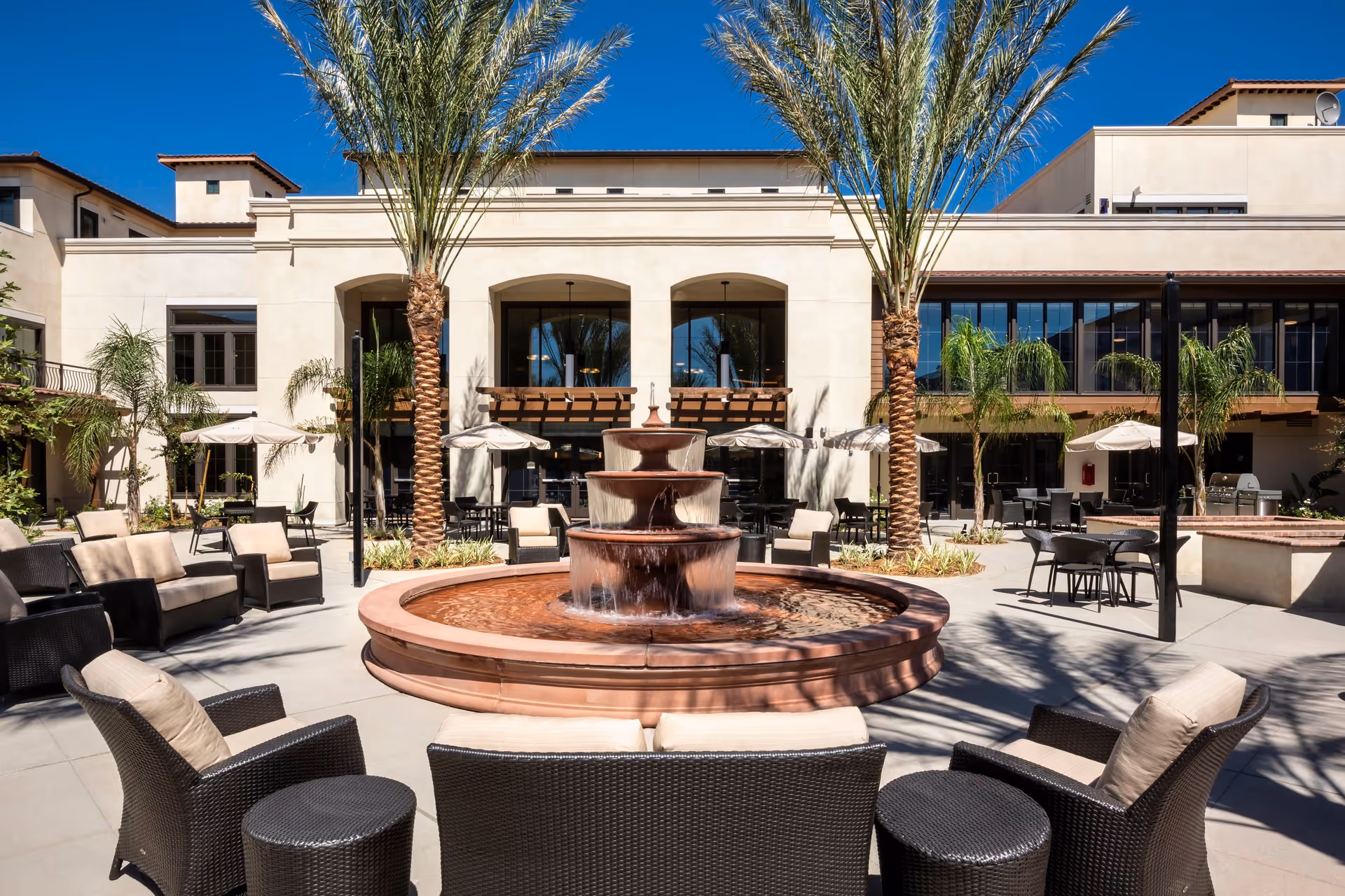 Outdoor courtyard area at Merrill Gardens at Rancho Cucamonga featuring a large tiered water fountain in the center, surrounded by cushioned wicker chairs and small tables. Tall palm trees and umbrellas provide shade, with a beige building and large windows in the background under a clear blue sky.