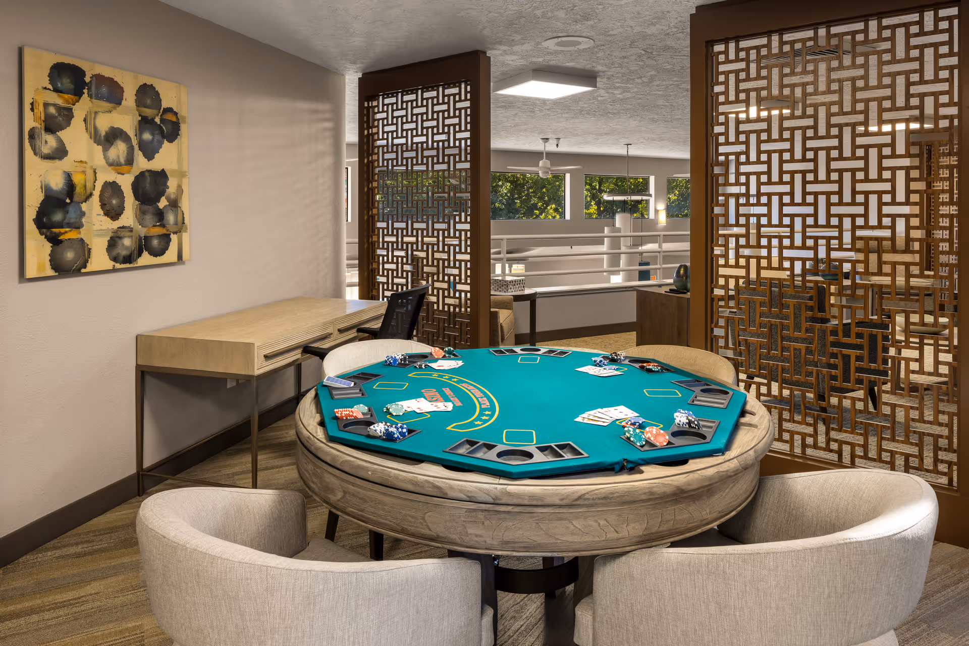 A cozy interior room featuring a round poker table with playing cards and poker chips set up for a game. The table is surrounded by four cushioned chairs. Behind the table, there is a wooden desk with a black office chair. Decorative wooden lattice panels partially separate this space from an adjacent area with large windows letting in natural light.