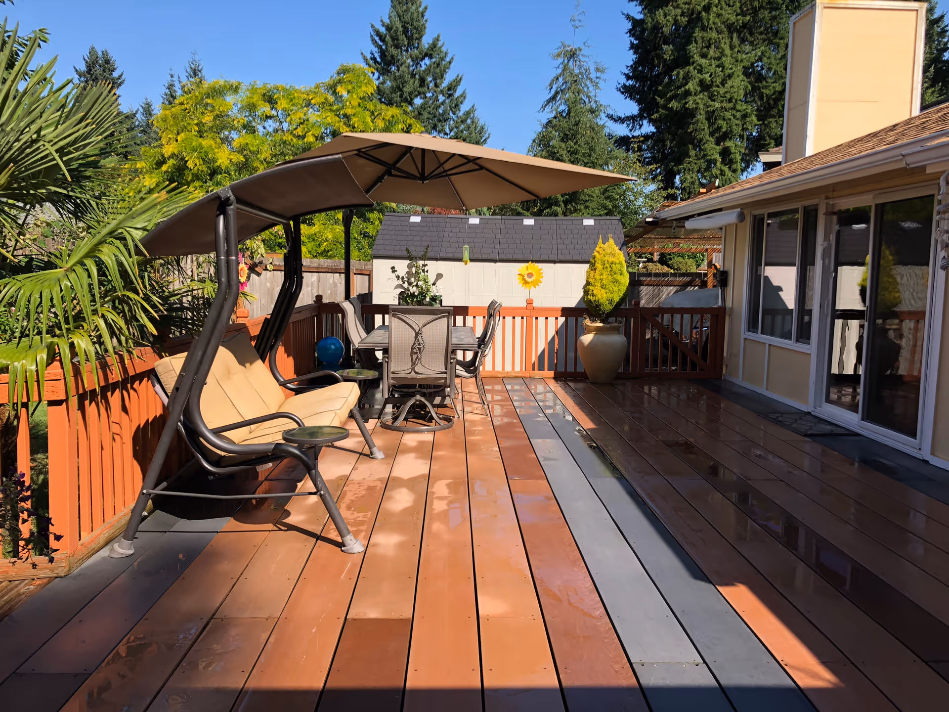 Outdoor wooden deck area with a cushioned swing bench under a canopy, a dining table with chairs, potted plants, and a small shed in the background surrounded by trees and a clear blue sky.