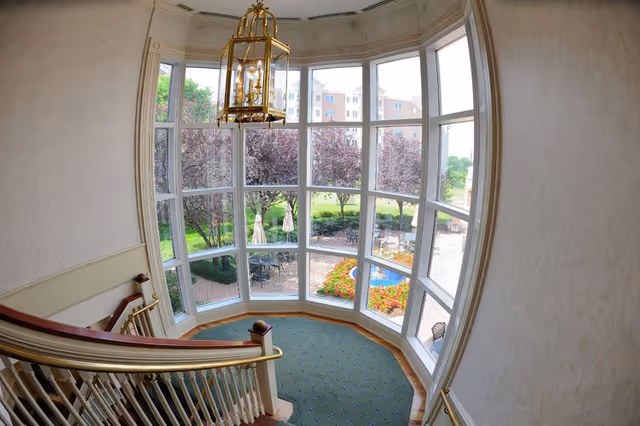 View from an indoor staircase landing with a large curved window overlooking an outdoor patio area with tables, chairs, umbrellas, flower beds, and trees. A gold chandelier hangs from the ceiling above the landing.