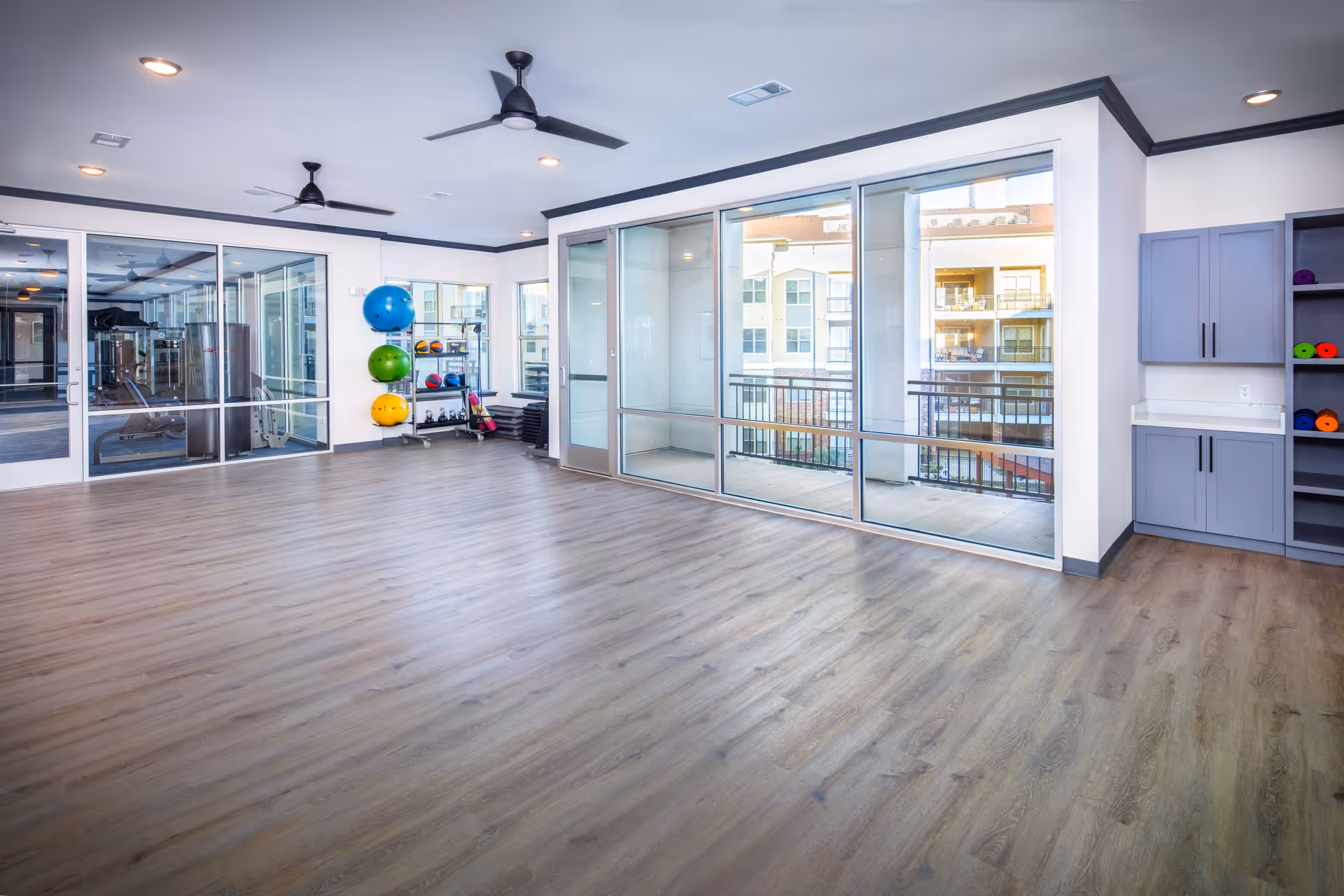 Bright multipurpose fitness studio with wood flooring, stacked exercise balls and floor-to-ceiling glass doors opening to a balcony.