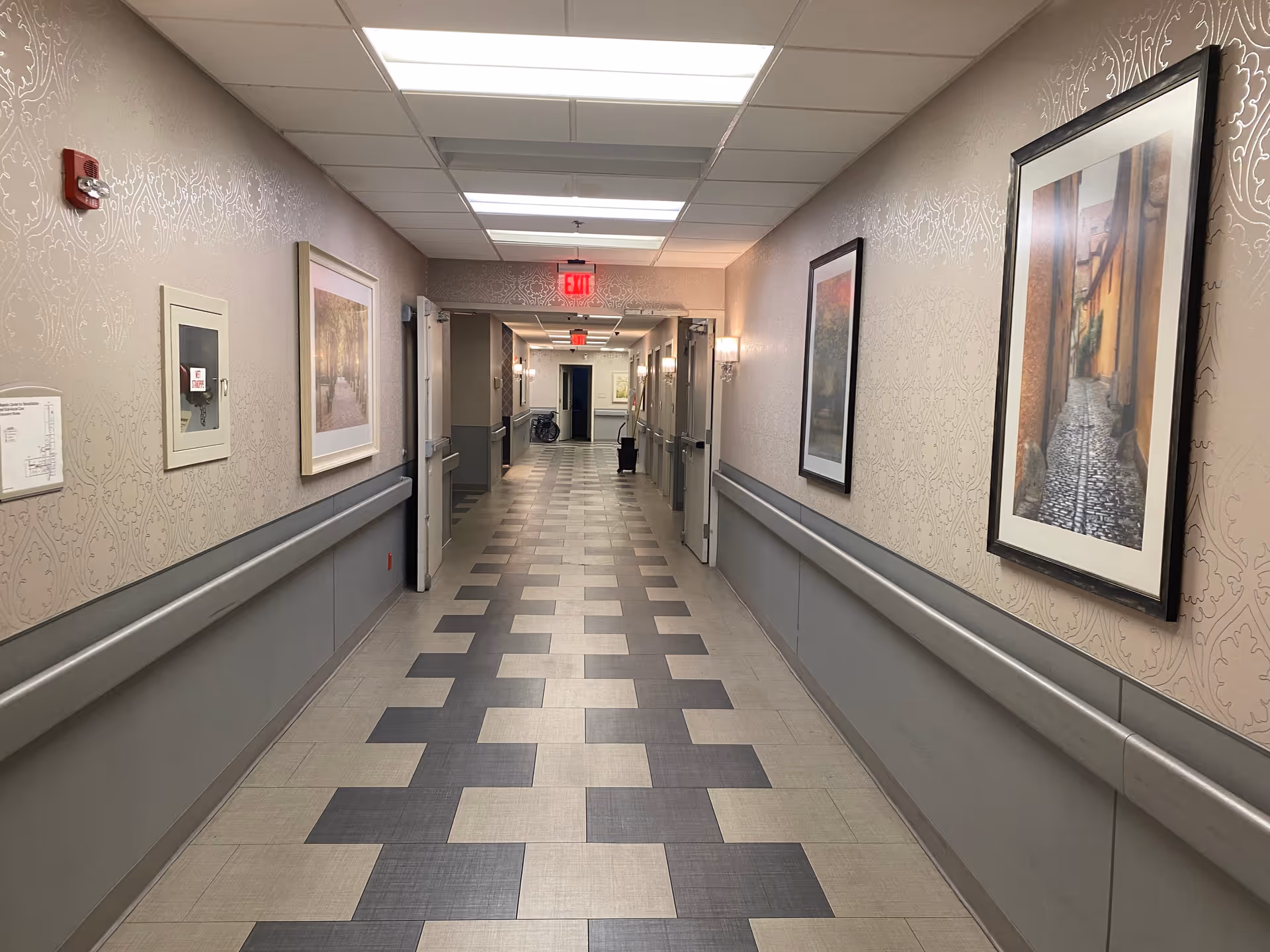 A long, well-lit hallway in a senior living facility with patterned tile flooring and beige walls adorned with framed artwork. The hallway has handrails on both sides and several doors leading to rooms. There are ceiling lights and exit signs visible at the end of the corridor.