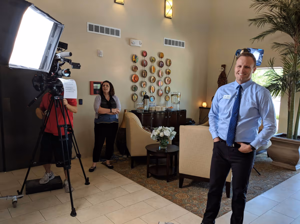 A cameraman with lighting and two people stand in a furnished senior living lobby with chairs, a side table with flowers, and decorative wall plates.