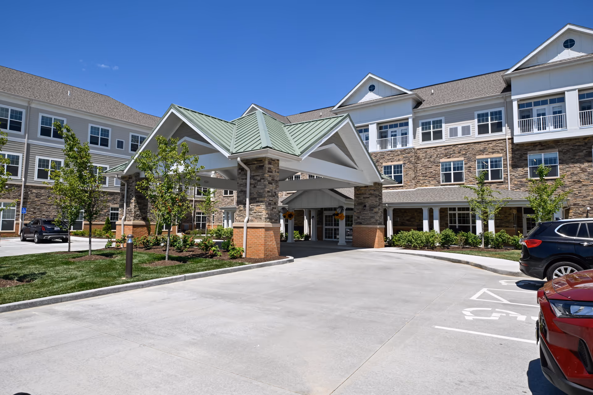 Exterior view of The Lumiere of Chesterfield senior living facility showing a large covered entrance with stone and brick pillars, surrounded by a well-maintained driveway, small trees, and parked cars under a clear blue sky.