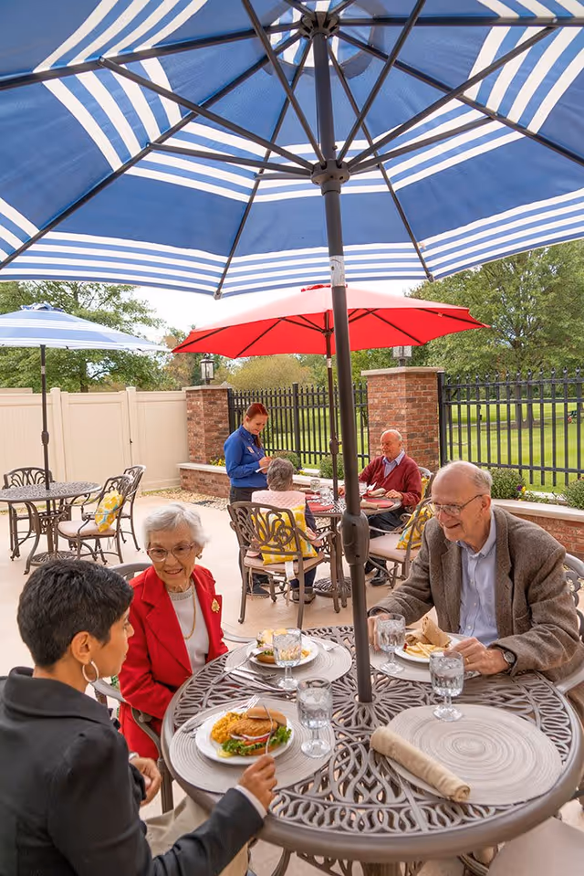 A group of elderly people and a caregiver enjoying a meal together at an outdoor patio with tables, chairs, and large umbrellas providing shade. The setting is surrounded by a fence and greenery.