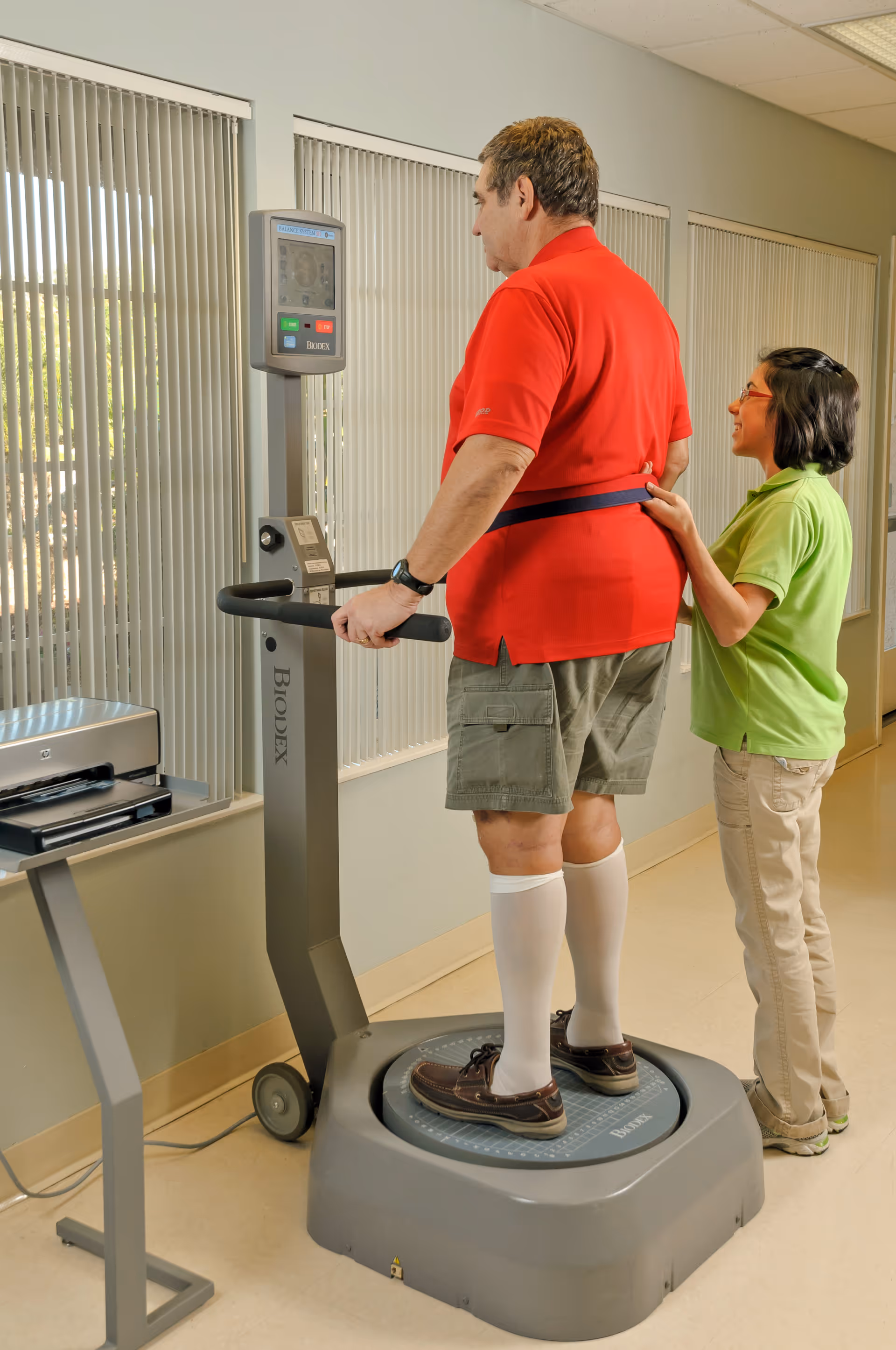 A man in a red shirt and khaki shorts stands on a Biodex balance testing machine while a woman in a green shirt assists him by holding a strap around his waist. They are in a room with vertical blinds covering the windows.