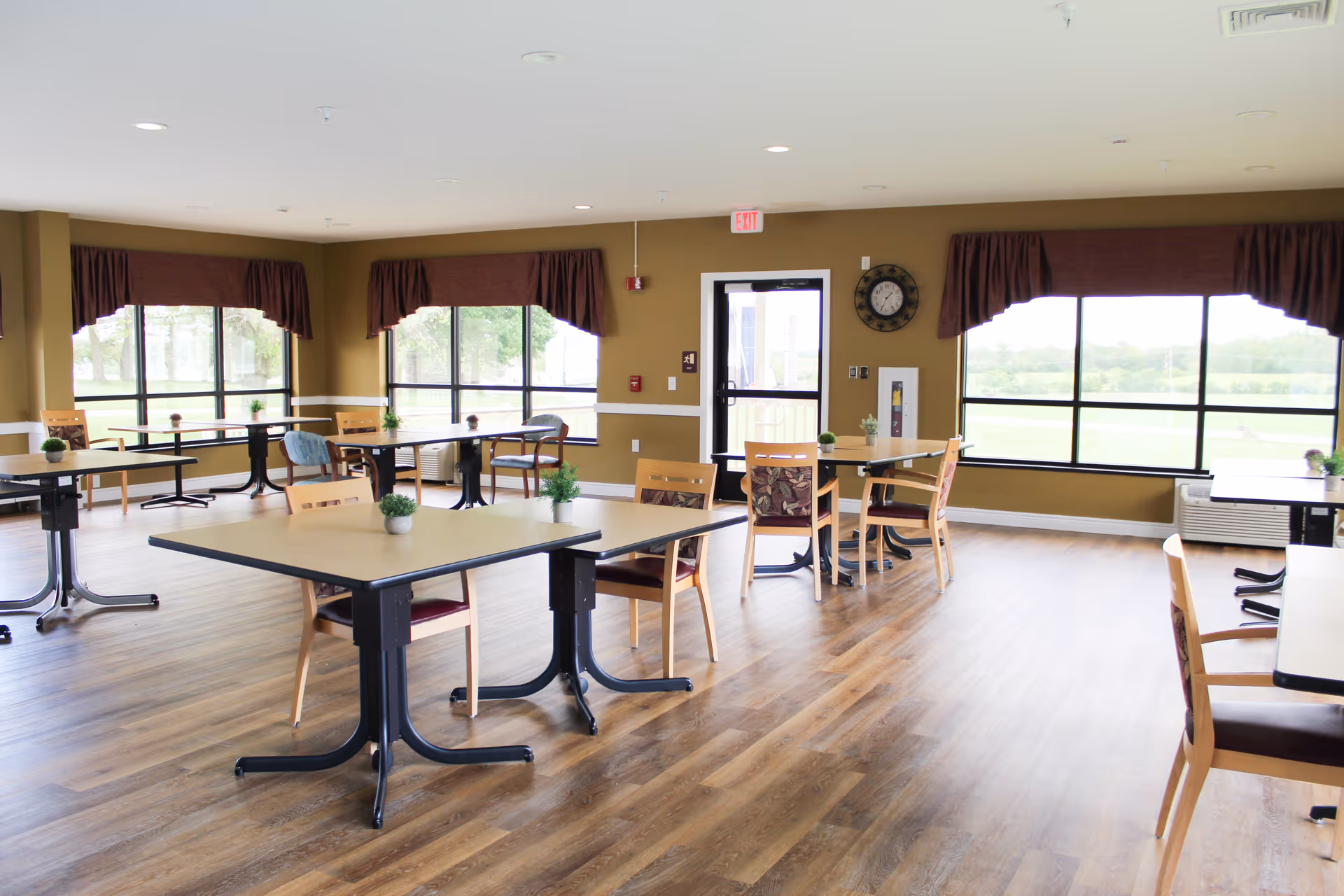 Bright communal dining area with multiple tables and chairs, large windows, and wood-look flooring.
