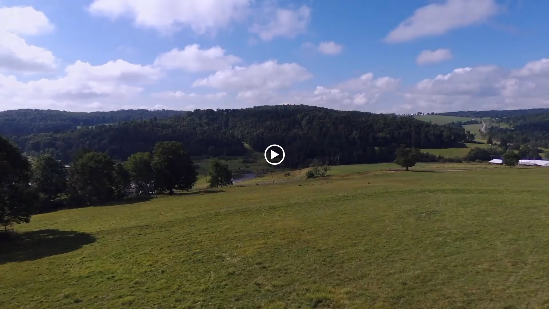 A wide aerial view of rolling green fields, scattered trees, and wooded hills under a partly cloudy sky.