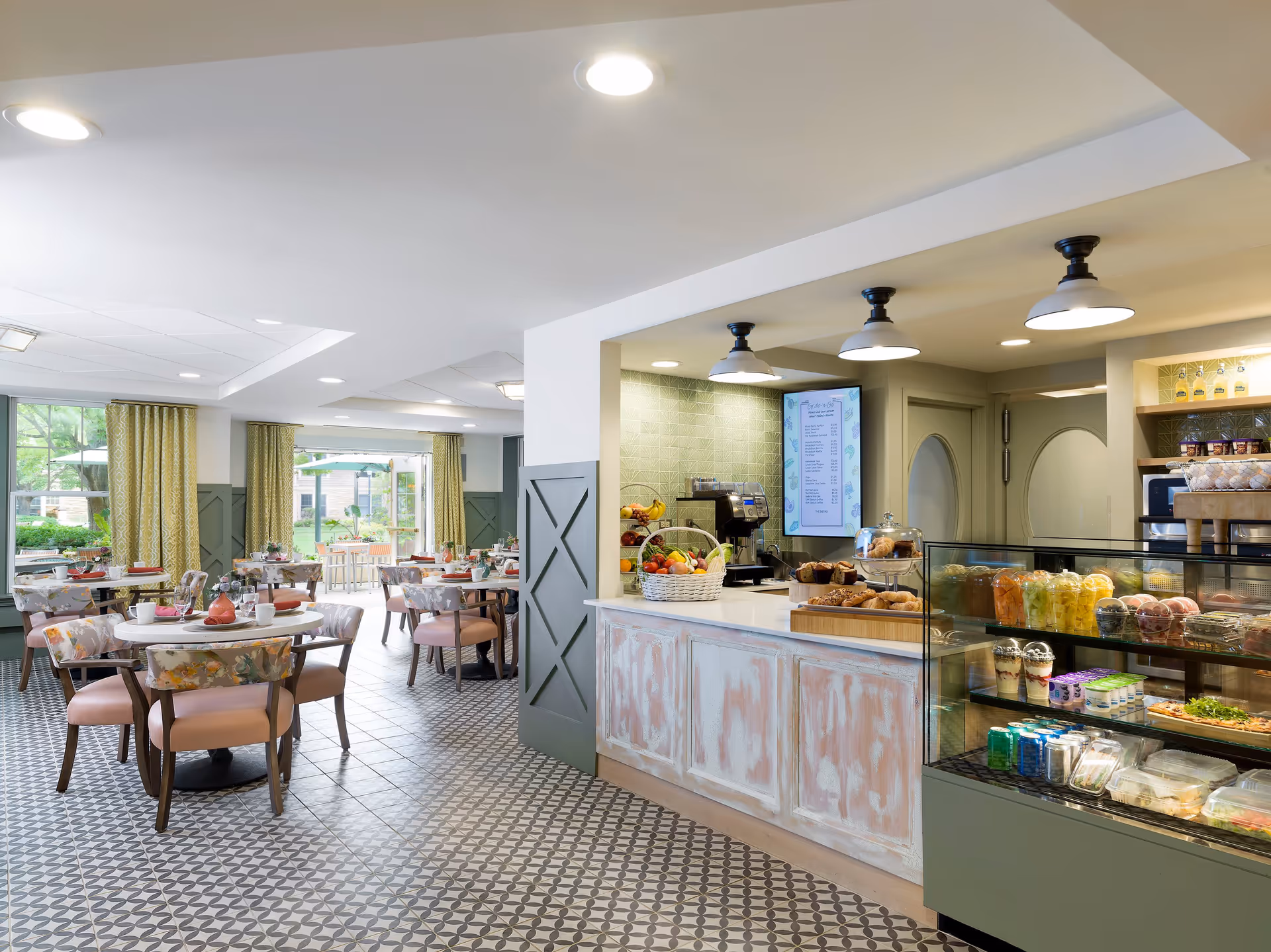 Bright and inviting dining area with round tables and cushioned chairs, set with cups and plates. To the right, a counter displays fresh fruit baskets, pastries, and a glass case with various packaged foods and beverages. Large windows with yellow curtains allow natural light to fill the space, and the floor features a patterned tile design.