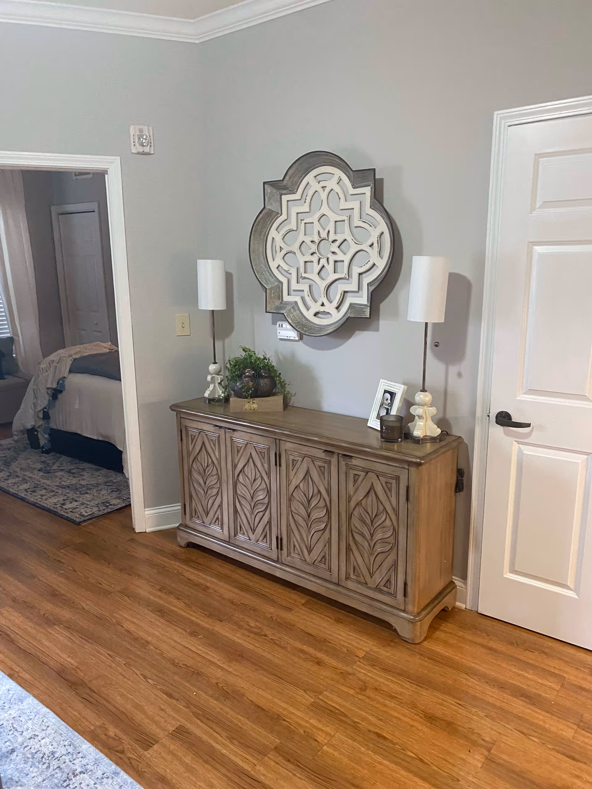 A hallway area with a wooden sideboard featuring carved leaf designs. On top of the sideboard are two tall lamps with white shades, a small plant, a framed photo, and a candle. Above the sideboard is a decorative wall mirror with an intricate geometric pattern. To the left, an open doorway reveals part of a bedroom with a bed, rug, and window with blinds. The floor is wood, and the walls are painted light gray.