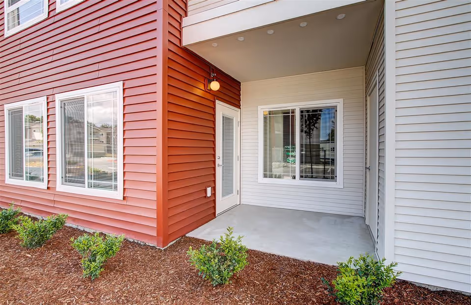 Covered outdoor patio area with concrete floor, adjacent to a building with red and white siding. There are windows with blinds and a door with a glass panel. Small green shrubs are planted in mulch along the edge of the patio.