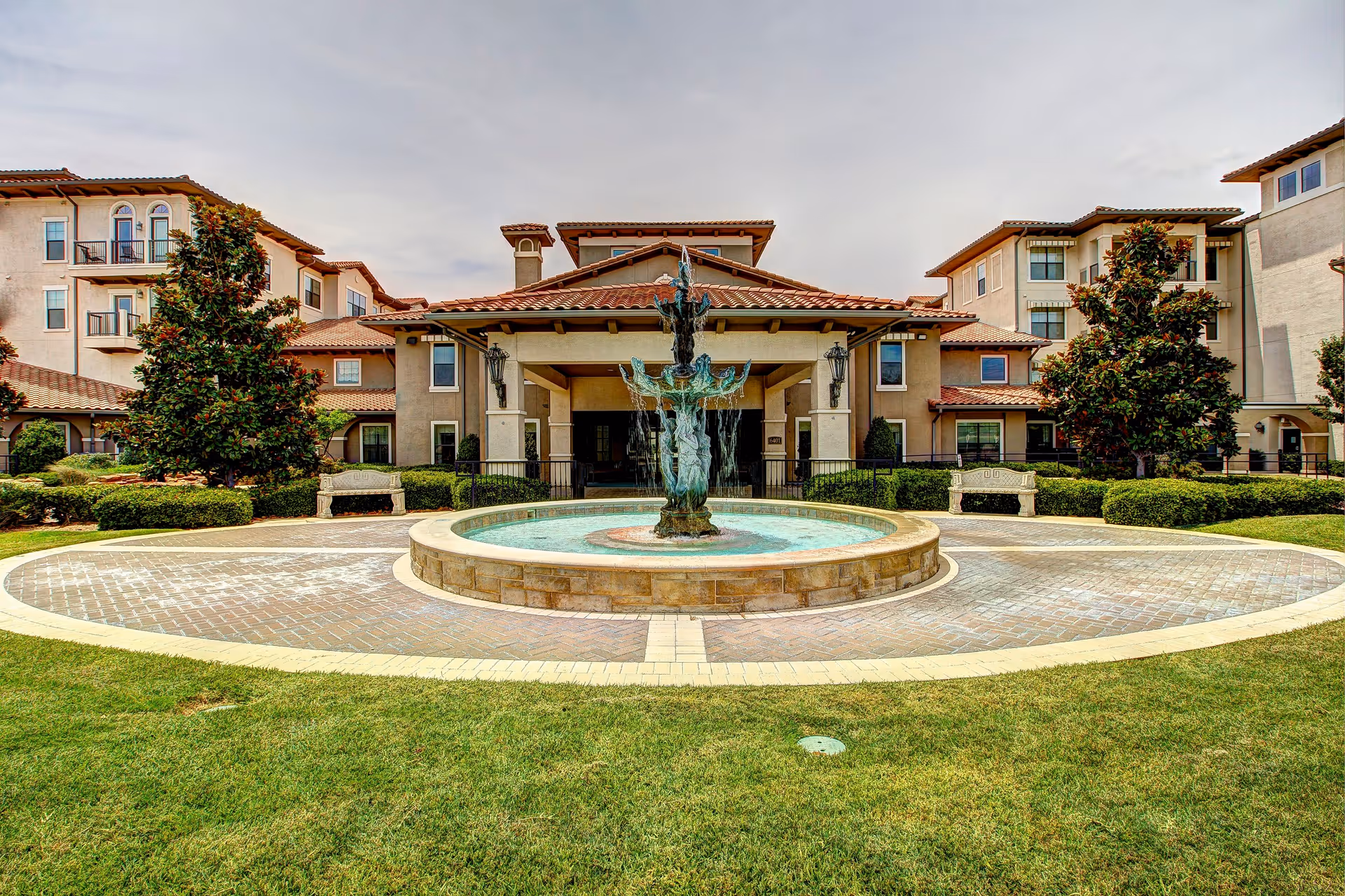 Exterior view of Conservatory At Plano senior living facility featuring a central water fountain surrounded by a circular paved area, green lawn, and multi-story buildings with balconies and tiled roofs under a cloudy sky.