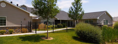 Exterior view of a single-story assisted living facility building with beige siding, a dark shingled roof, and multiple windows. The building is surrounded by a well-maintained lawn, trees, bushes, and a black metal fence enclosing a patio area.