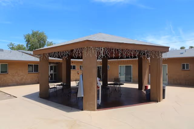 Outdoor covered patio area with a hexagonal roof supported by six columns, decorated with hanging string lights. Under the roof are several metal tables and chairs. Surrounding the patio is a single-story building with beige stucco walls and multiple windows and sliding glass doors. The sky is clear and blue.