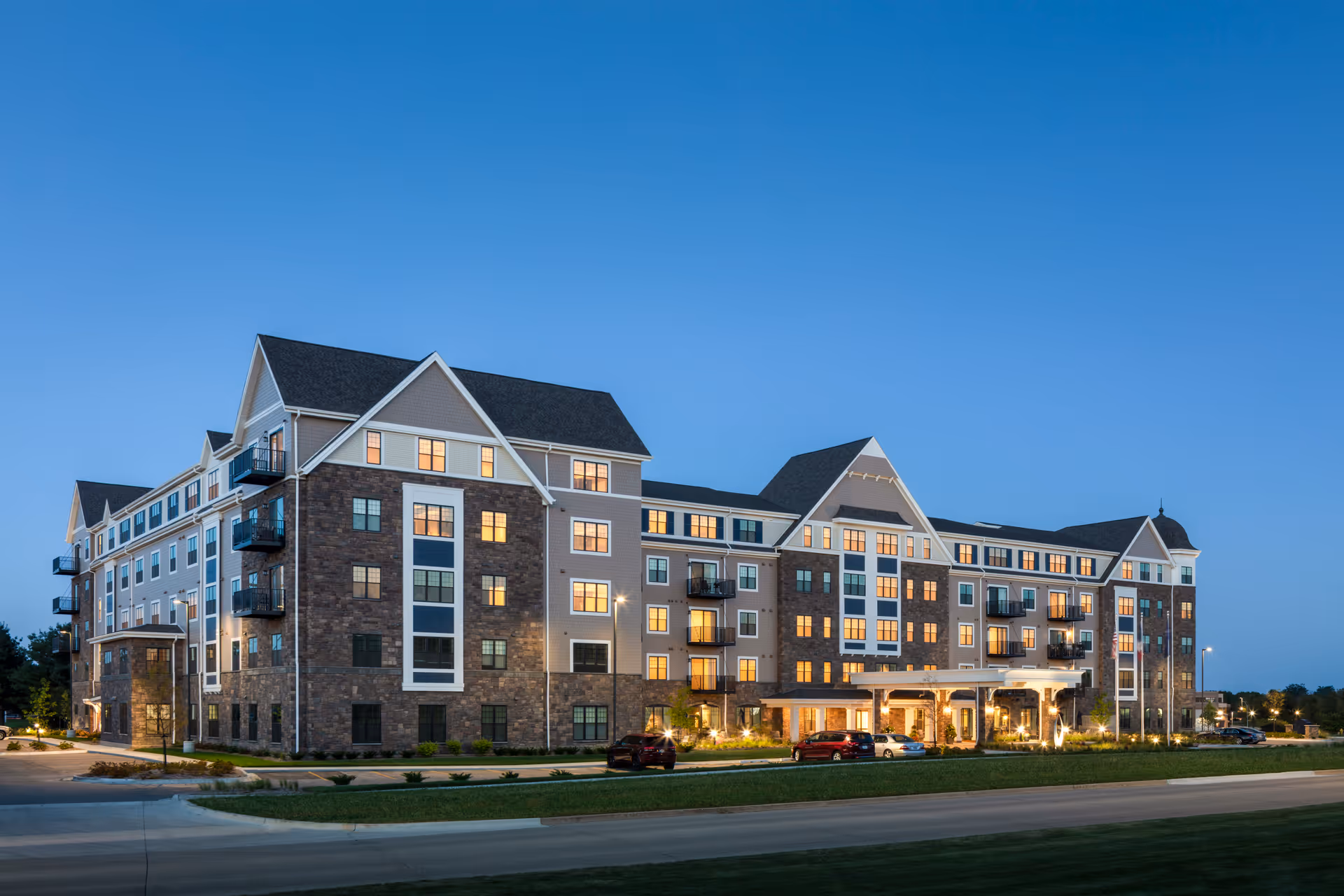 Exterior view of a large, multi-story senior living facility building named Grand Living At Tower Place during twilight with lights glowing from the windows and cars parked in front.