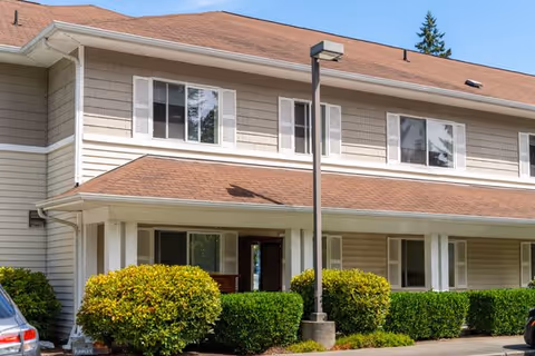 Exterior view of a two-story residential building with beige siding and a brown roof. The building has multiple windows with white shutters and a covered entrance supported by white columns. There are well-maintained green bushes and a streetlamp in front of the building under a clear blue sky.