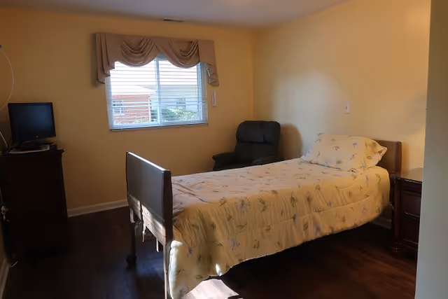 Sunlit bedroom with a single bed, a recliner chair, a small TV on a dresser, and a window with a valance.