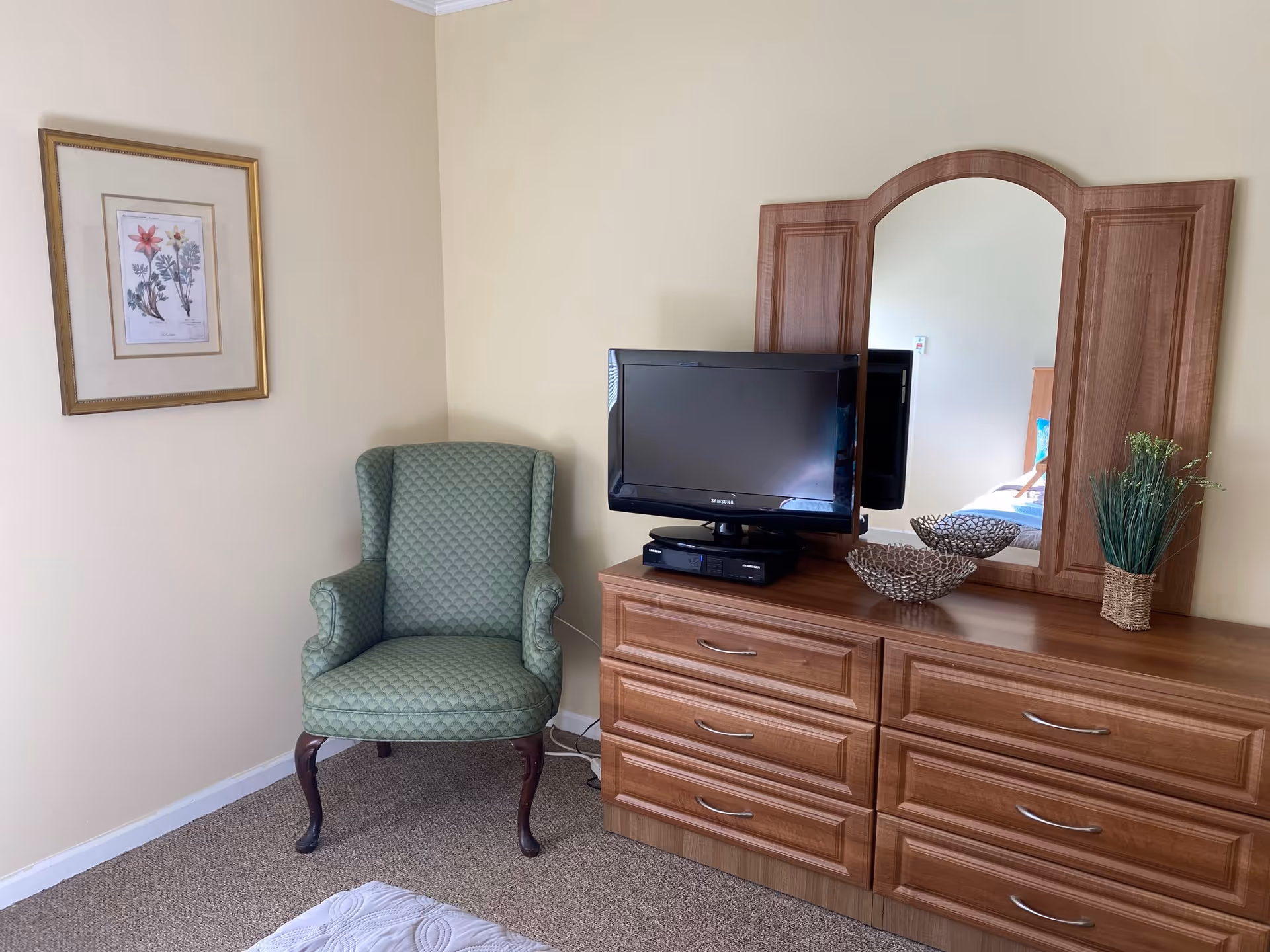 A corner of a bedroom featuring a green upholstered armchair with wooden legs, a wooden dresser with six drawers, a flat-screen Samsung TV on top of the dresser, a decorative bowl, a small plant in a woven basket, and a large wooden-framed mirror. A framed floral artwork hangs on the wall above the armchair.