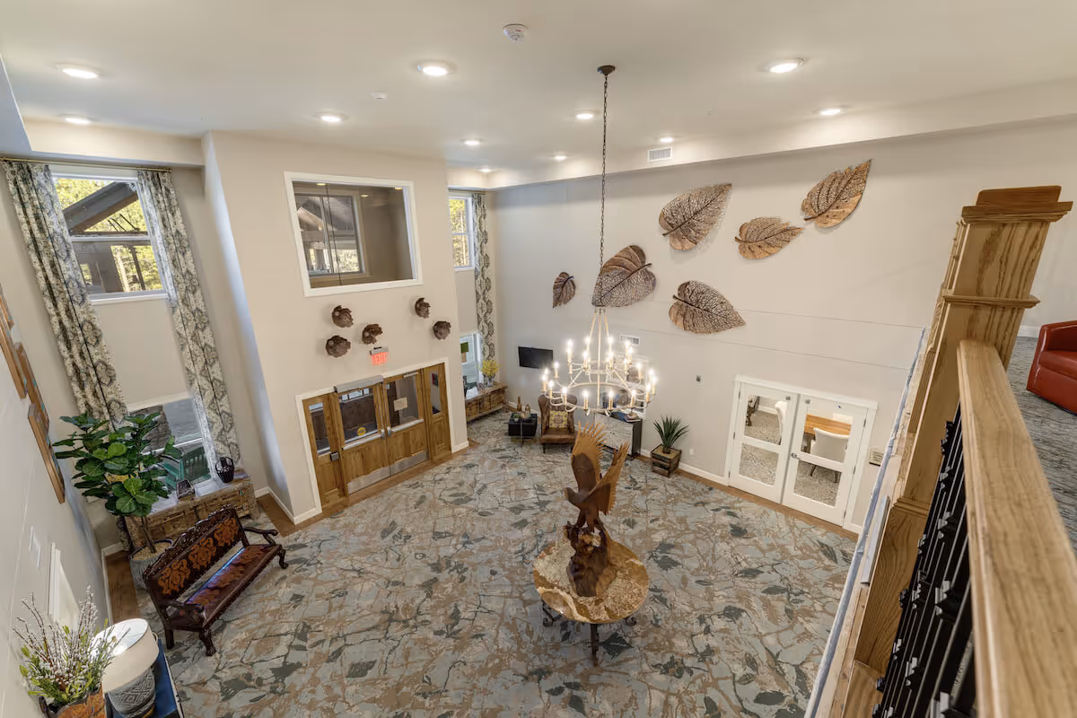 Spacious senior living facility lobby with a high ceiling, decorative leaf wall art, a chandelier, and a wooden eagle sculpture on a round table. The room features large windows with patterned curtains, wooden double doors, a bench, and plants, creating a welcoming atmosphere.