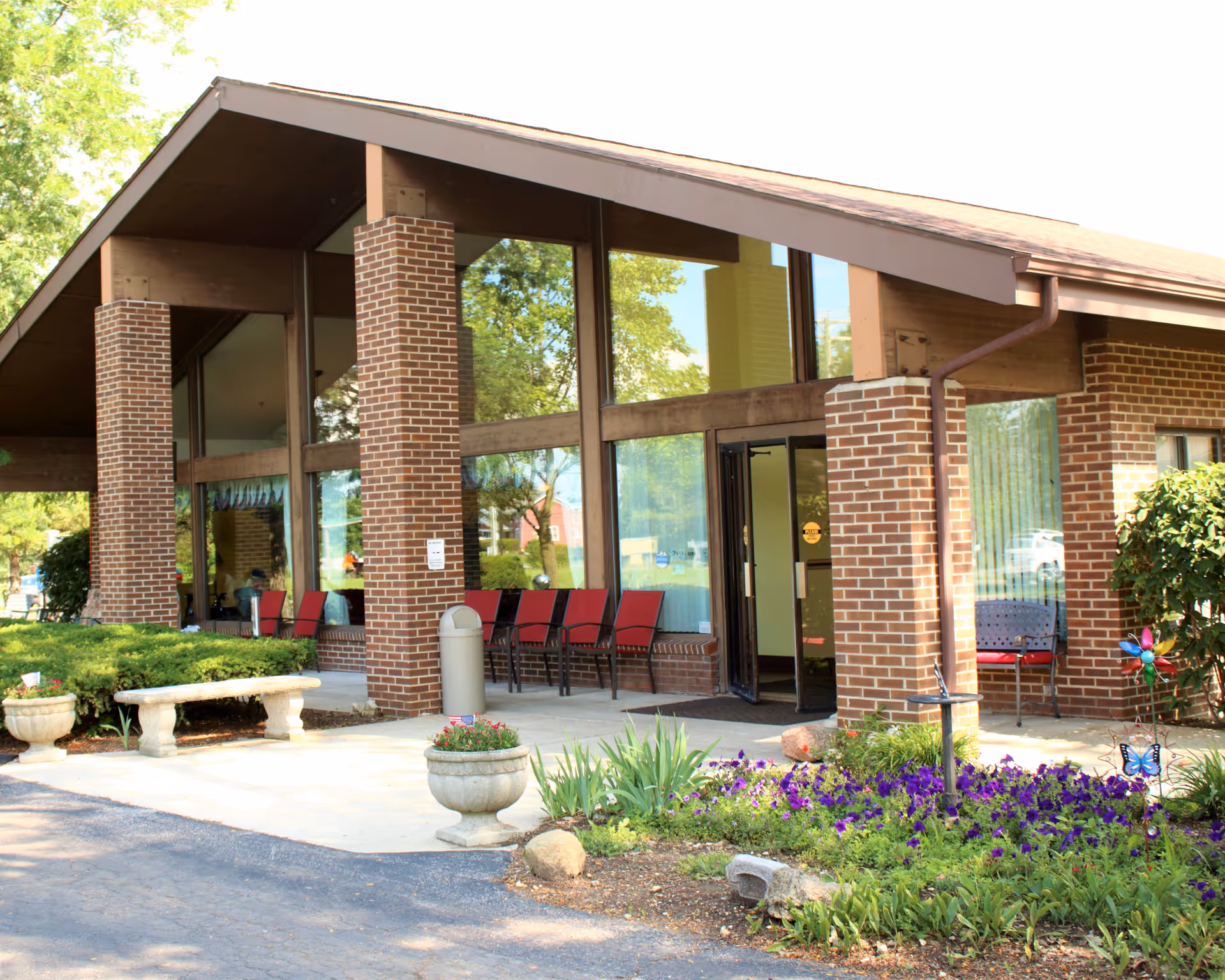 Front entrance of Autumnwood Care Center with brick columns, large windows, outdoor chairs, planters, and flower beds.