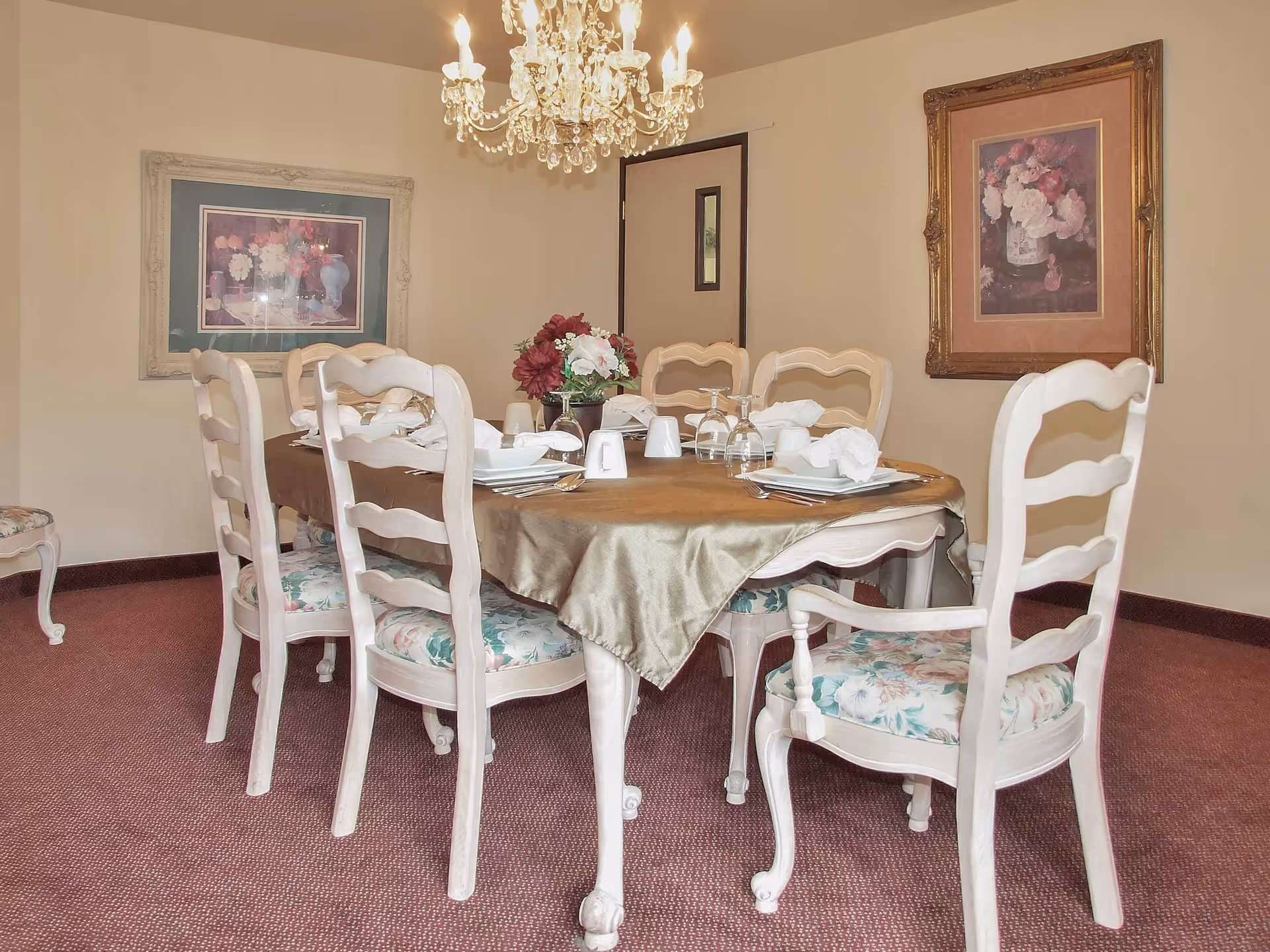 A formal dining room with a round table set for dinner, upholstered floral chairs, a floral centerpiece and a crystal chandelier.