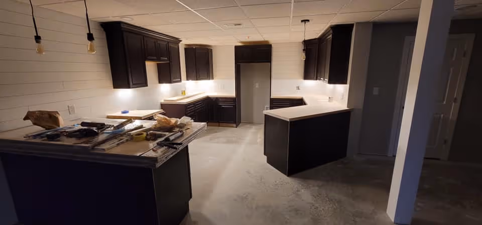 Interior view of a kitchen under renovation with dark wood cabinets, white countertops, and tools and materials placed on the central island. The floor is unfinished concrete, and the walls are light-colored with a shiplap design. Pendant lights hang from the ceiling.