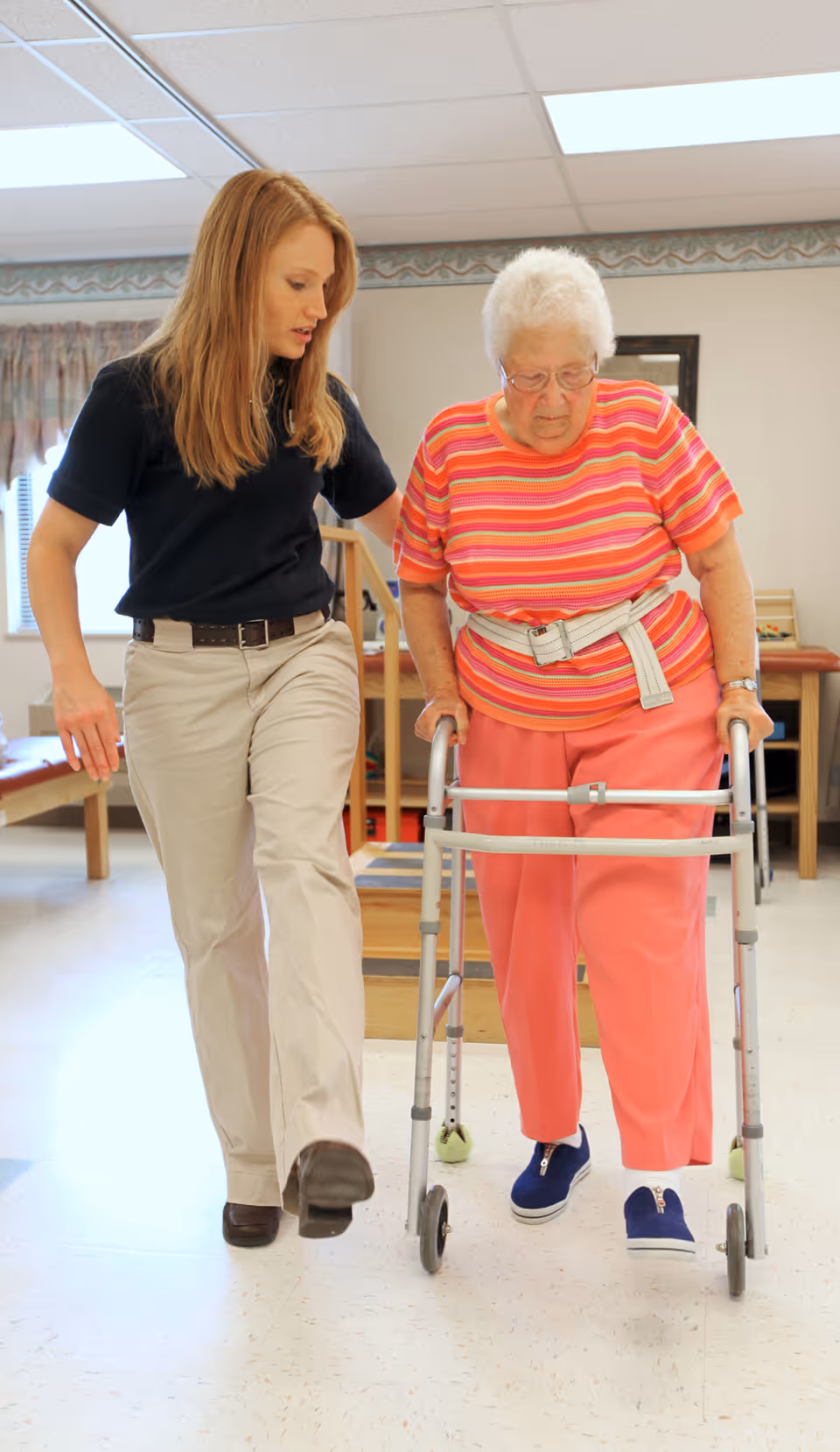 A young female caregiver assists an elderly woman using a walker as they walk together indoors in a well-lit room with exercise equipment in the background.