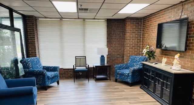 A cozy sitting area with three blue patterned armchairs and one wooden chair arranged around a small wooden side table with a blue lamp. The room has brick walls, a large window with white blinds, a wooden floor, and a black cabinet with decorative figurines and a flat-screen TV mounted above it.