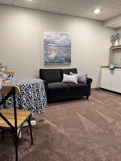 A cozy interior room featuring a black loveseat with two decorative pillows, one with the word 'smile' on it. To the left is a table covered with a black and white patterned tablecloth and a wooden chair. On the wall above the loveseat hangs a painting of a seascape with sailboats. To the right, there is a white cabinet with a wooden countertop and a small plant on a shelf above it. The room has beige walls and carpeted flooring.
