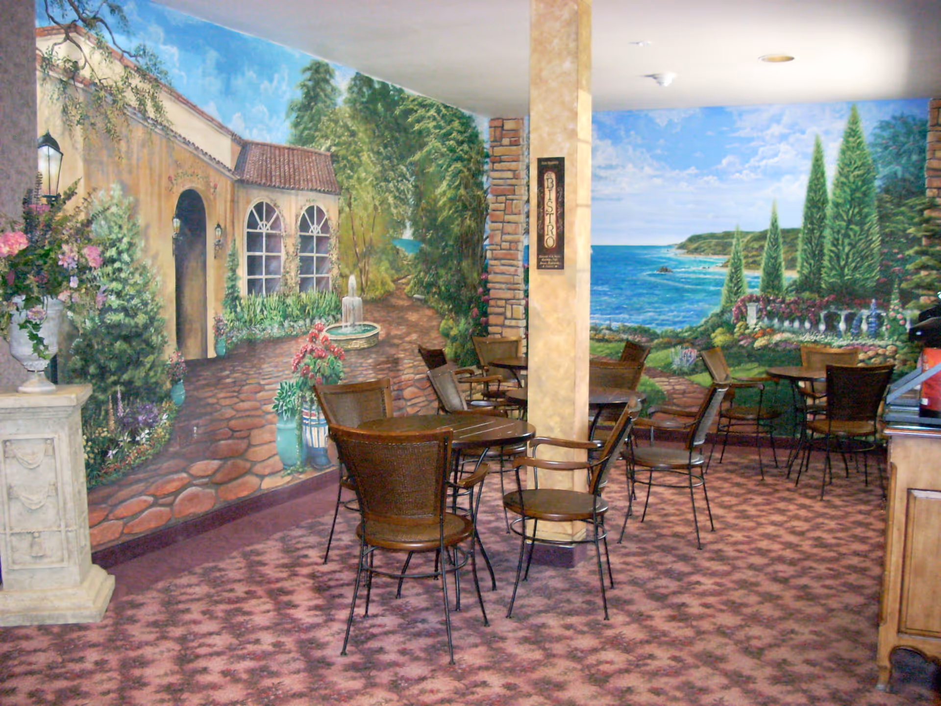 Dining area with several round tables and chairs set against colorful seaside and courtyard wall murals.