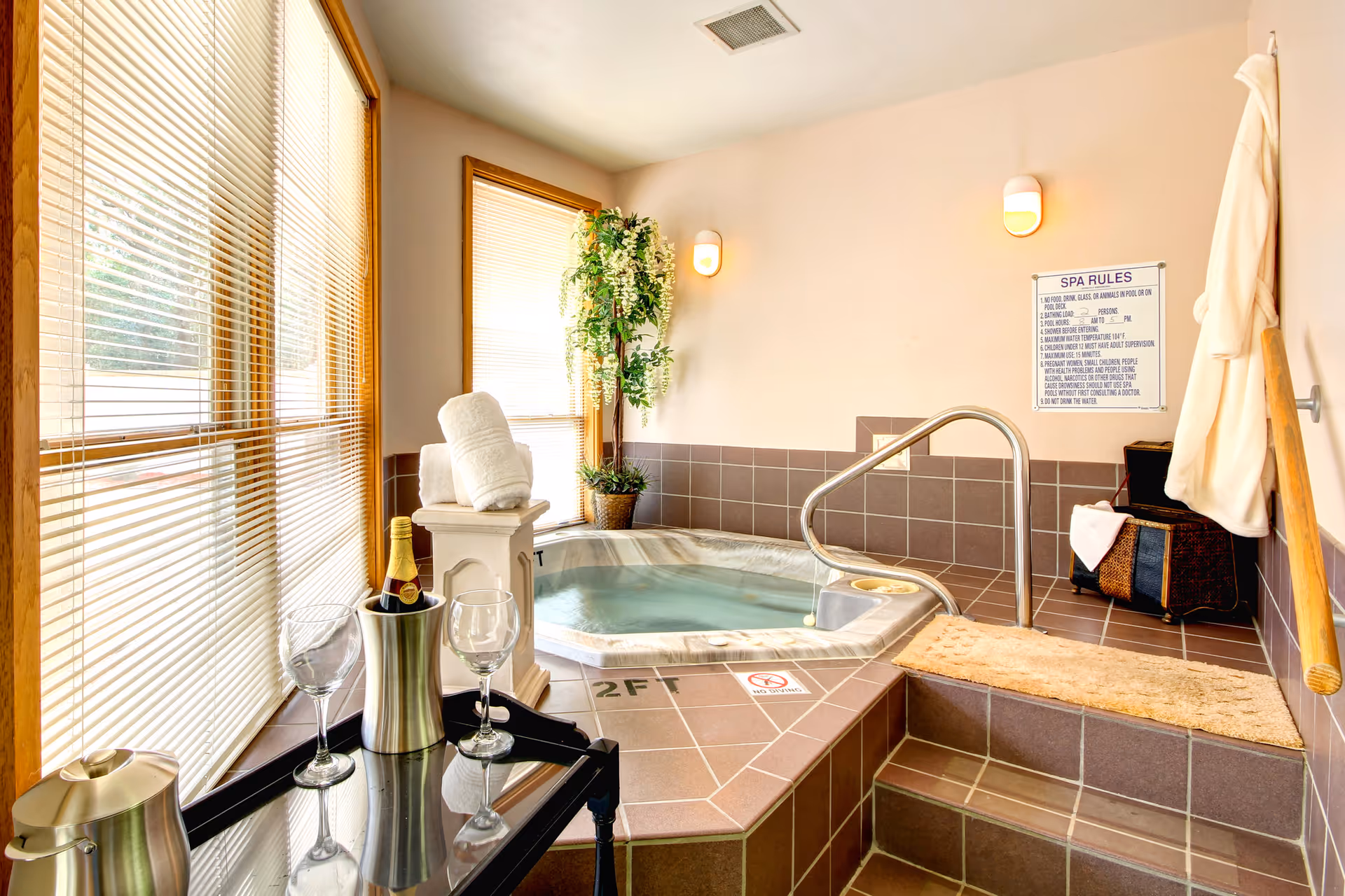 Tiled indoor spa room with a small hot tub and handrail, rolled towels, a potted plant, and a cart with wine and glasses by large windows.