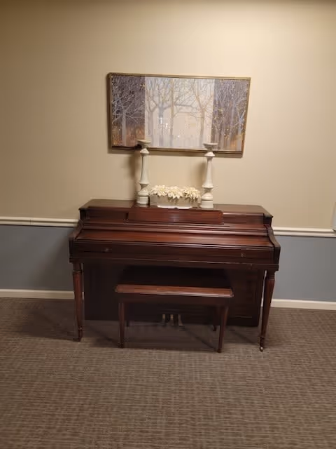 A wooden upright piano with a matching bench placed on a carpeted floor against a beige and gray wall. On top of the piano are two white candlesticks and a decorative flower arrangement. Above the piano hangs a framed painting depicting a forest scene with trees.