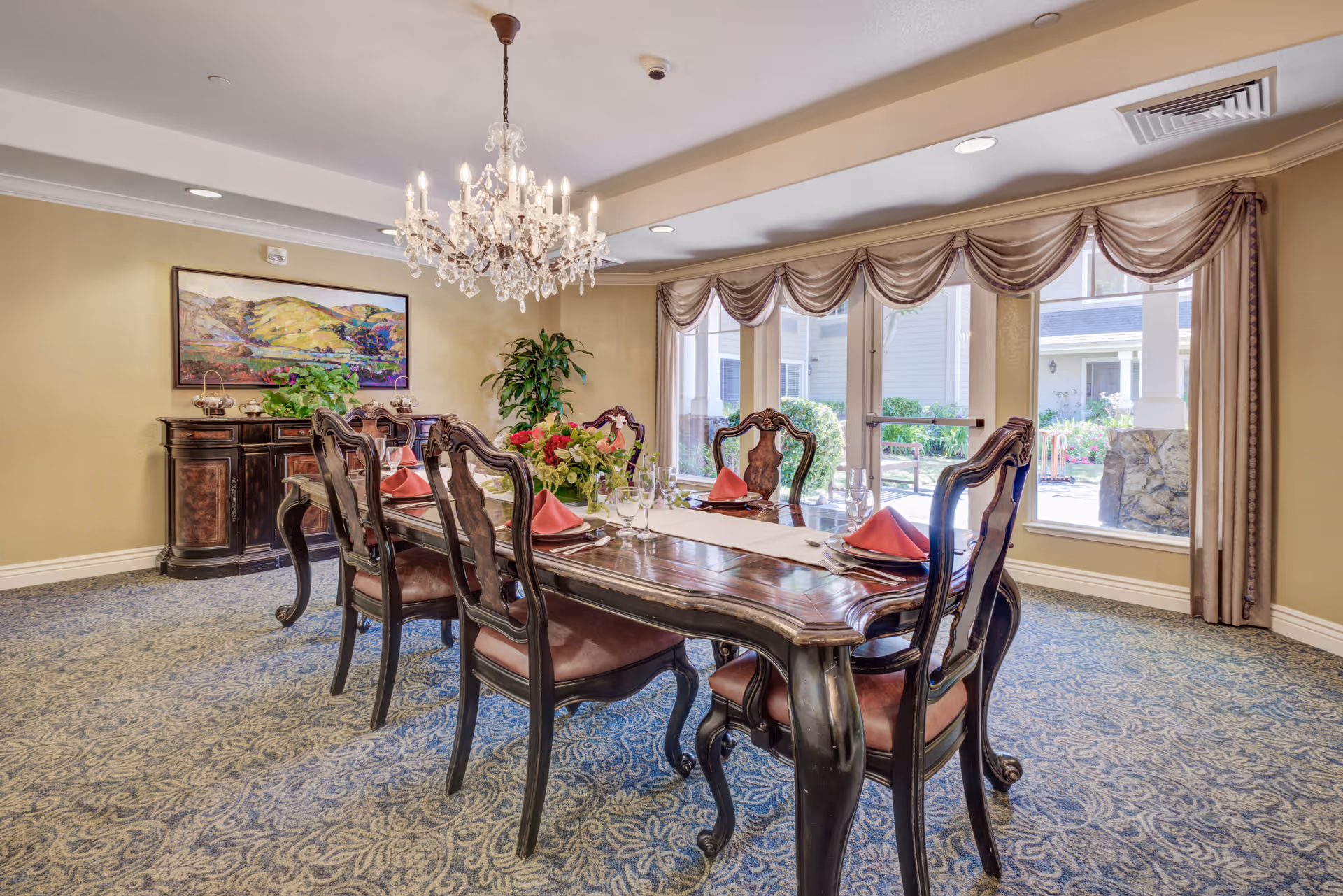 Elegant dining room with a long wooden table set with red napkins and glassware. Six ornate wooden chairs surround the table. A crystal chandelier hangs above, and large windows with draped curtains allow natural light to fill the room. A sideboard with plants and a landscape painting is visible against the wall.