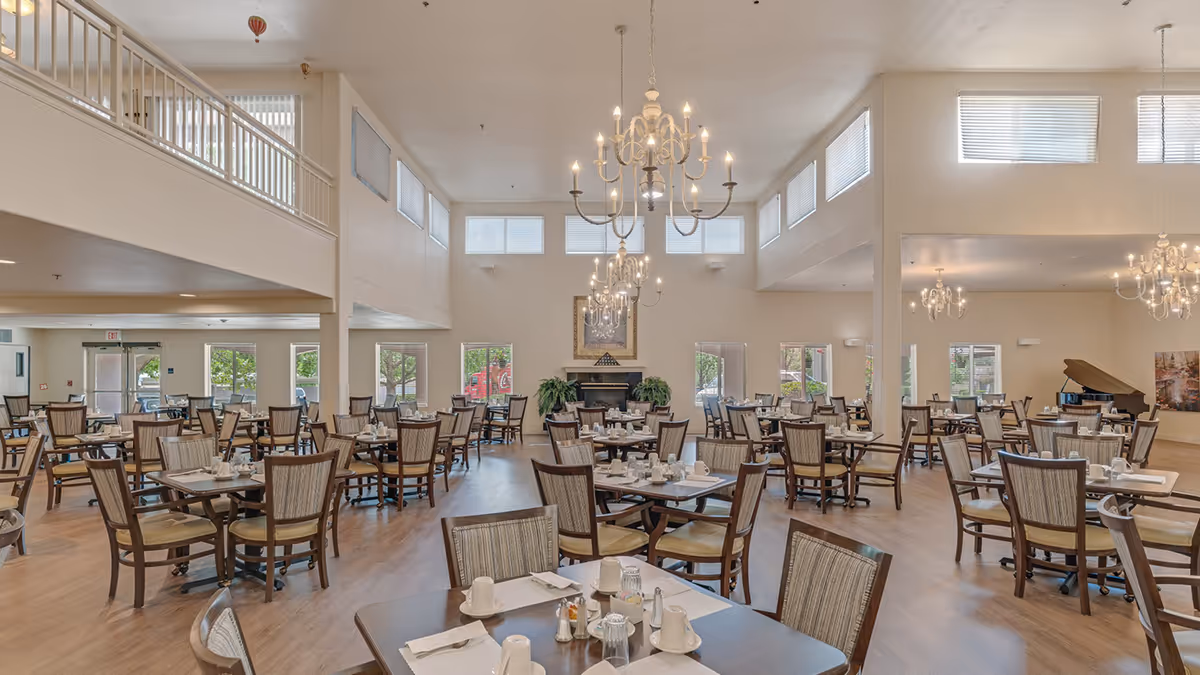 Spacious dining room with multiple wooden tables and chairs arranged neatly. Each table is set with white cups, glasses, napkins, and silverware. The room features high ceilings with several elegant chandeliers hanging down, large windows allowing natural light, and a fireplace with plants on either side. A grand piano is visible in the background.