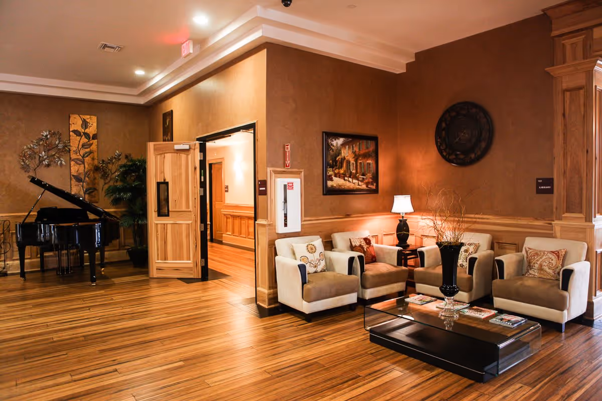 A cozy sitting area in an assisted living facility with four beige armchairs arranged around a glass coffee table with magazines. A black vase with decorative branches is on the table. The walls are painted brown with wooden paneling, and there is a table lamp, a painting, and a large decorative clock on the walls. To the left, there is a black grand piano and some plants near an open wooden door leading to a hallway.