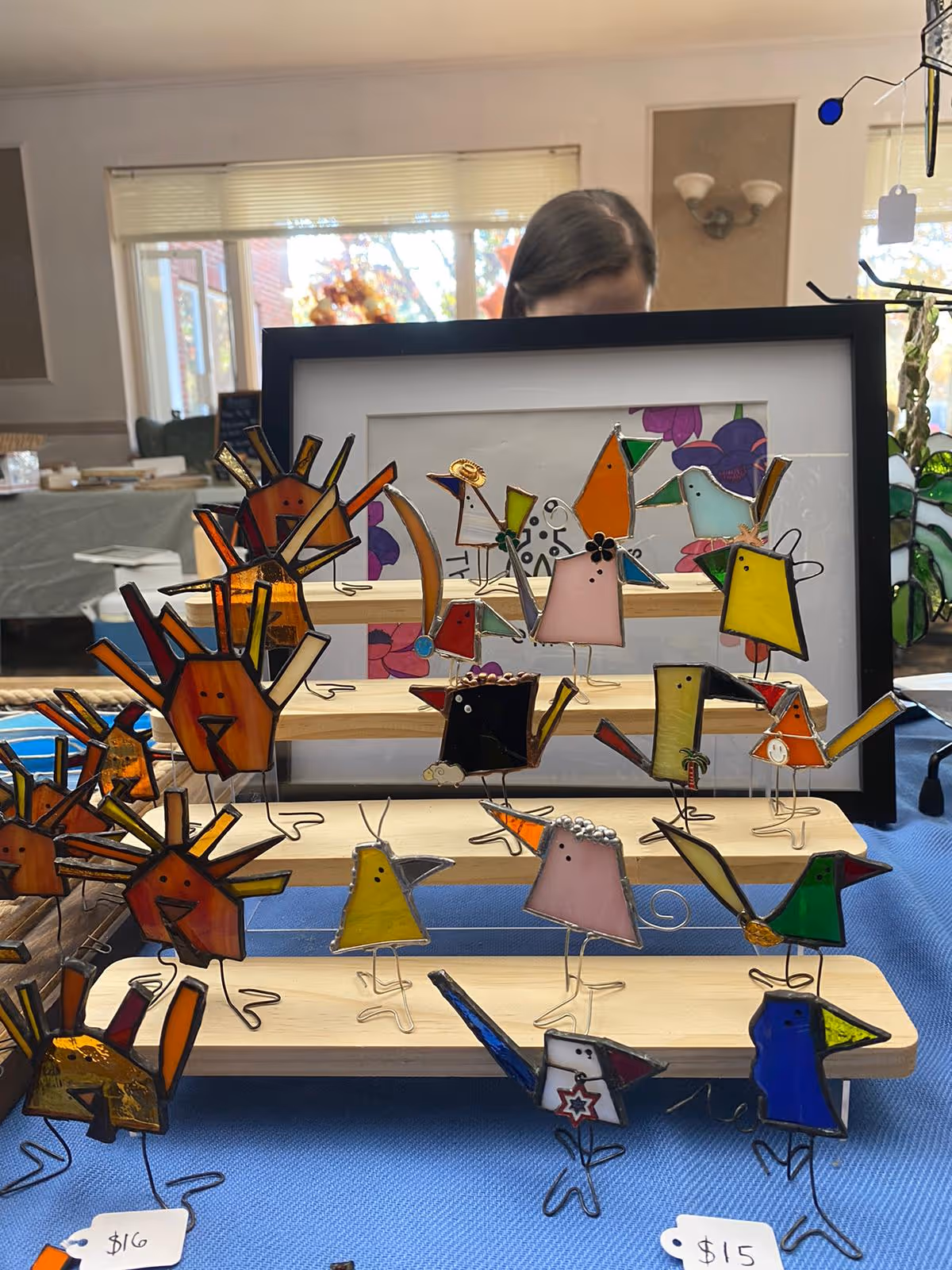 A display of colorful stained glass bird and turkey figurines arranged on wooden stands on a blue tablecloth inside a room with large windows and a person partially visible behind the display.