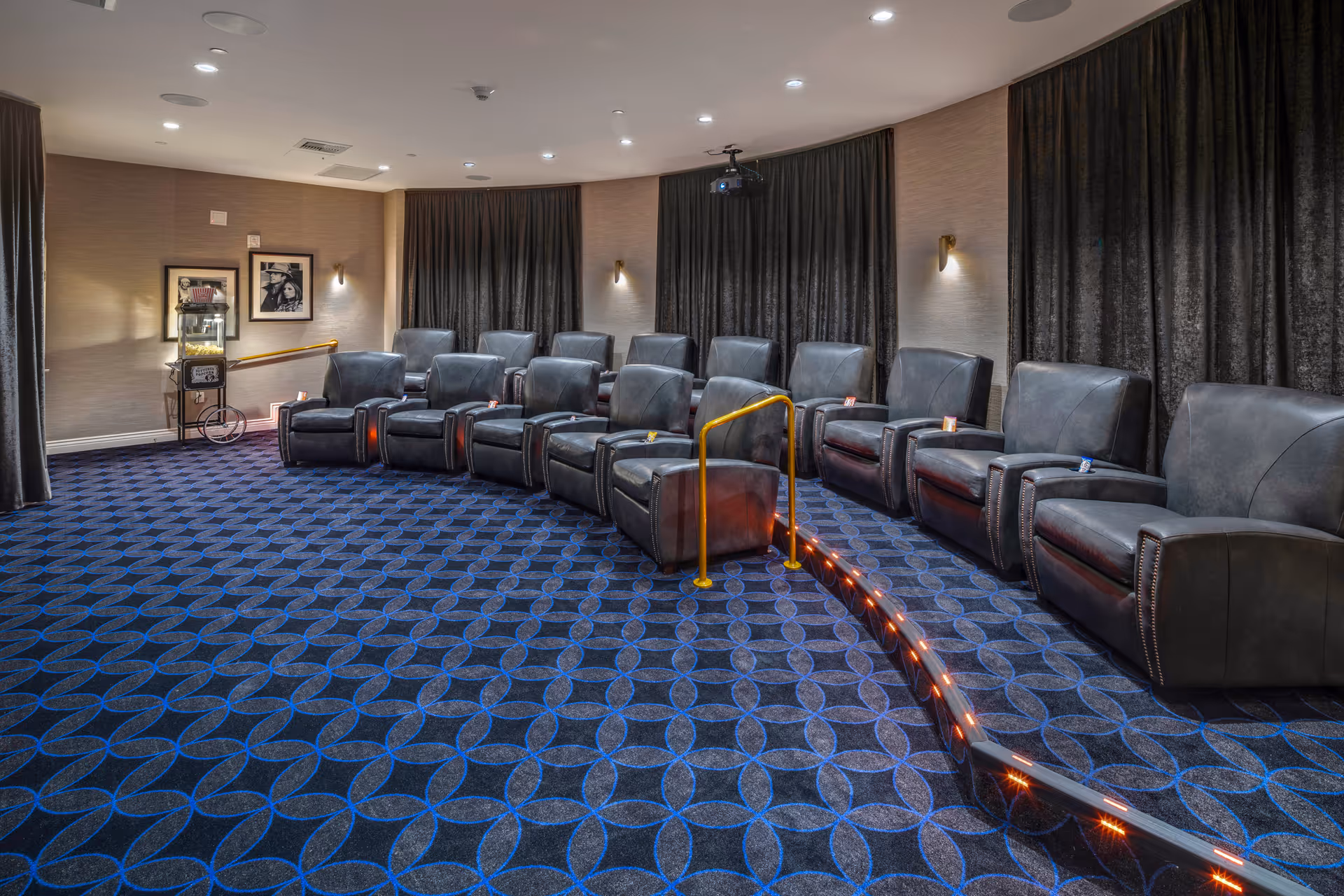 Small private movie theater with leather recliner seats arranged on a curved riser, patterned carpet, projector, and a popcorn machine.