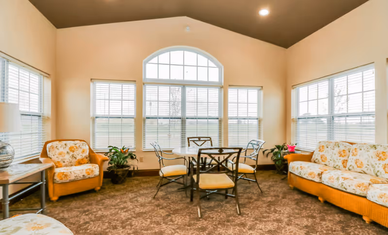 Sunlit seating area with floral-upholstered chairs and a sofa arranged around a round table in front of large multi-pane windows.